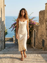 Load image into Gallery viewer, THE TARA handcrafted leather strappy sandal in camel on model  stone steps flower shrub and water backdrop — LUXUL COLLECTIVE